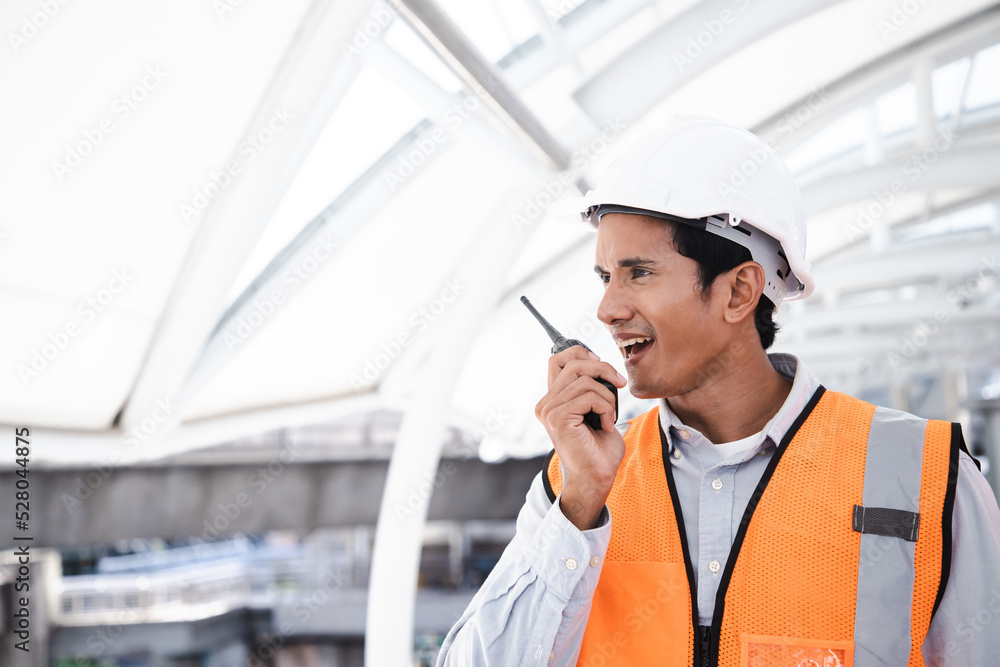 Portrait of handsome engineering man using walkie talkie and wear hardhat in front of construction outside work station. Back view of contractor on background of modern buildings. Portrait of handsome engineering man using walkie talkie and wear hardhat in front of construction outside work station. Back view of contractor on background of modern buildings.