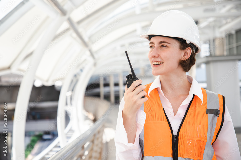 Portrait of beautiful engineering woman using walkie talkie and wear hardhat in front of construction outside work station. Back view of contractor on background of modern buildings. Portrait of beautiful engineering woman using walkie talkie and wear hardhat in front of construction outside work station. Back view of contractor on background of modern buildings.