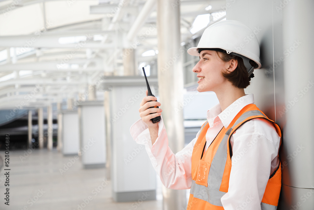 Portrait of beautiful engineering woman using walkie talkie and wear hardhat in front of construction outside work station. Back view of contractor on background of modern buildings. Portrait of beautiful engineering woman using walkie talkie and wear hardhat in front of construction outside work station. Back view of contractor on background of modern buildings.