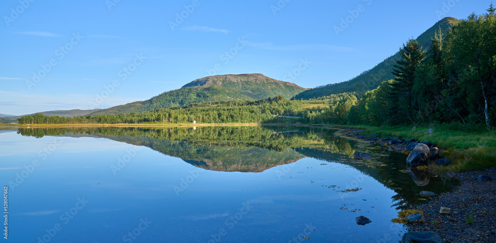 Mountain range in the Lofoten Island, Norway with a symmetry reflection in the water of a lake. Natural mirror. Sunny morning without clouds in the sky. Summer vacation in Lofoten.