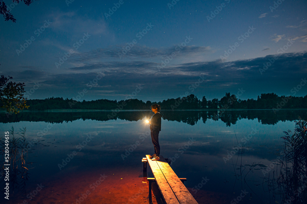 a young girl with a flashlight stands on the shore of a forest lake, as the lighthouse illuminates the way to the shore. Waiting for a guy from fishing. evening comes,the first stars appear in the sky - obrazy, fototapety, plakaty 
