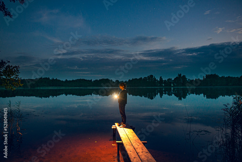 Fotografija a young girl with a flashlight stands on the shore of a forest lake, as the lighthouse illuminates the way to the shore