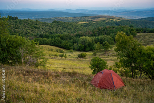 Dramatic landscape with a red tent in the foreground and a vast forest stretching in the distance. Camping in wild hill and mountain areas.