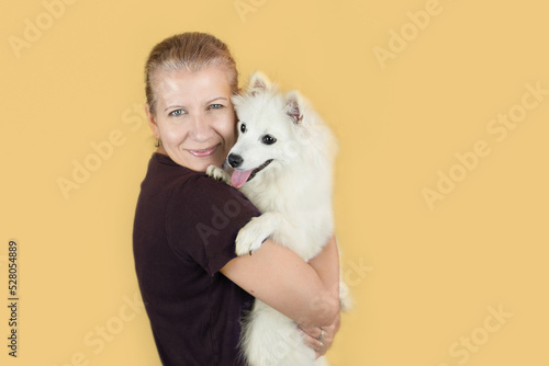 Fototapeta Naklejka Na Ścianę i Meble -  Beautiful joyful mature woman hugs her reliable friend - white spitz dog, woman is happy to have someone to talk to, woman with dog in her arms on yellow background