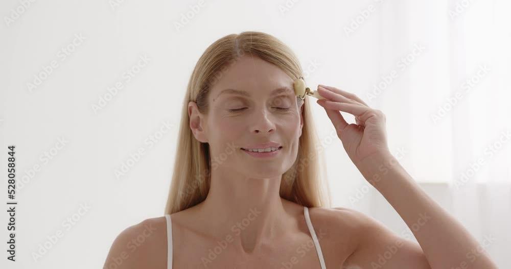 Gorgeous woman massaging face skin using facial trendy jade quartz roller device Gorgeous woman massaging face skin using facial trendy jade quartz roller device