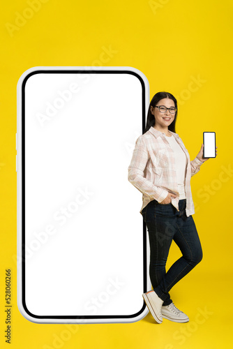 Looking at camera asian woman leaned on big smartphone with white screen showing phone in her hand wearing casual isolated on yellow background. Copy space on right, phone mock up