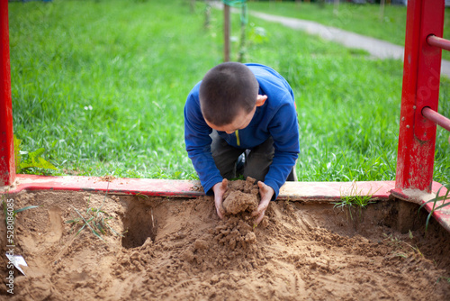 Child plays in sandbox. Boy in blue jacket on playground. Preschooler touches sand with his hands. Walk in summer in yard.