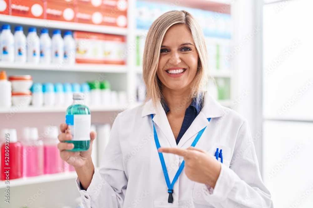 Blonde woman working at pharmacy drugstore holding syrup smiling happy pointing with hand and finger Blonde woman working at pharmacy drugstore holding syrup smiling happy pointing with hand and finger