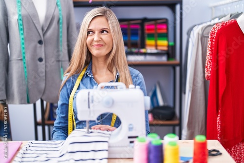 Blonde woman dressmaker designer using sew machine smiling looking to the side and staring away thinking.