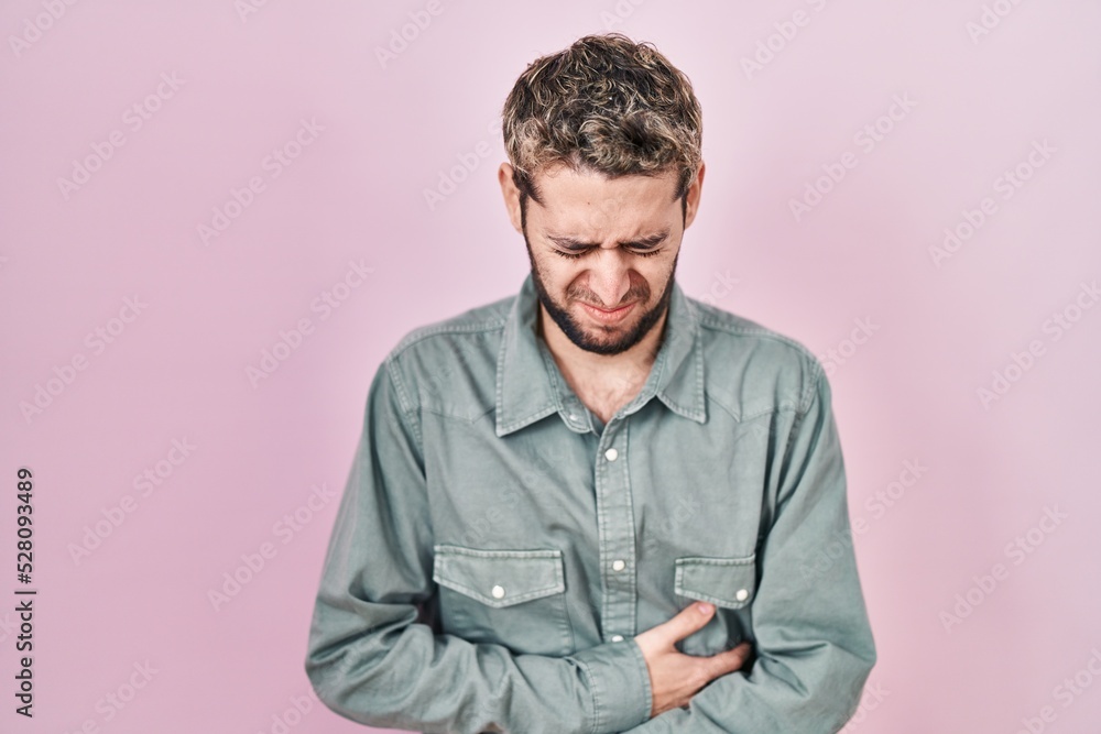 Hispanic man with beard standing over pink background with hand on stomach because nausea, painful disease feeling unwell. ache concept. Hispanic man with beard standing over pink background with hand on stomach because nausea, painful disease feeling unwell. ache concept.