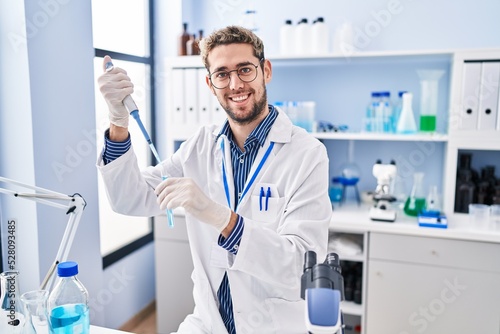 Young man scientist smiling confident pouring liquid on test tube at laboratory