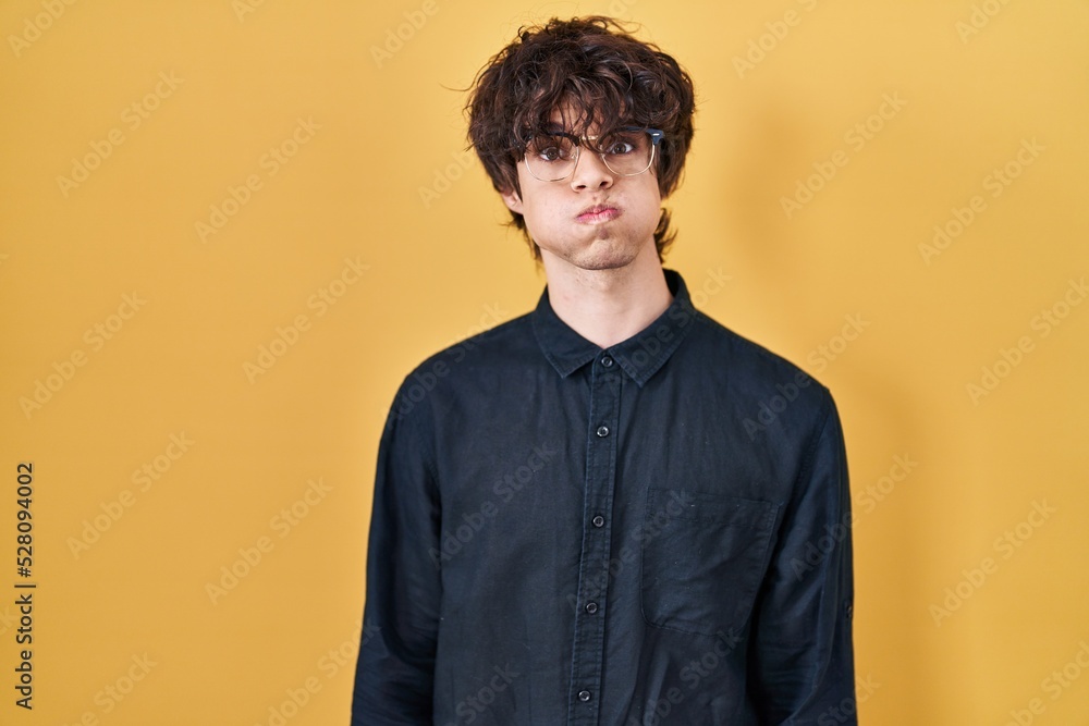 Young man wearing glasses over yellow background puffing cheeks with funny face. mouth inflated with air, crazy expression. Young man wearing glasses over yellow background puffing cheeks with funny face. mouth inflated with air, crazy expression.