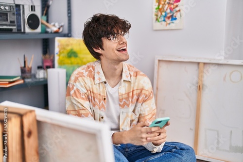 Young hispanic man artist smiling confident using smartphone at art studio