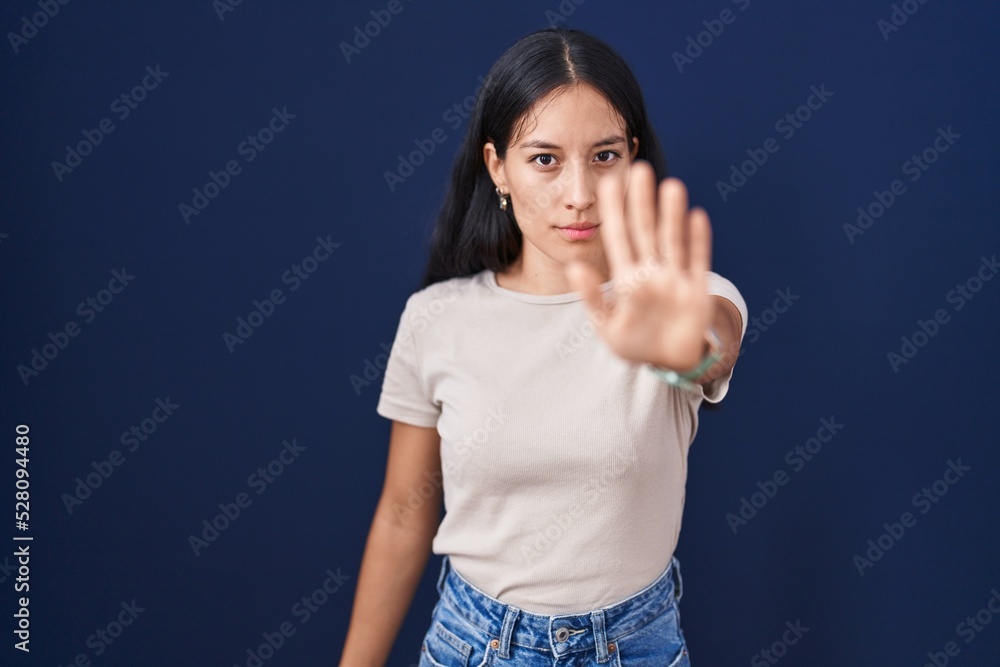 Fototapeta premium Young hispanic woman standing over blue background doing stop sing with palm of the hand. warning expression with negative and serious gesture on the face.