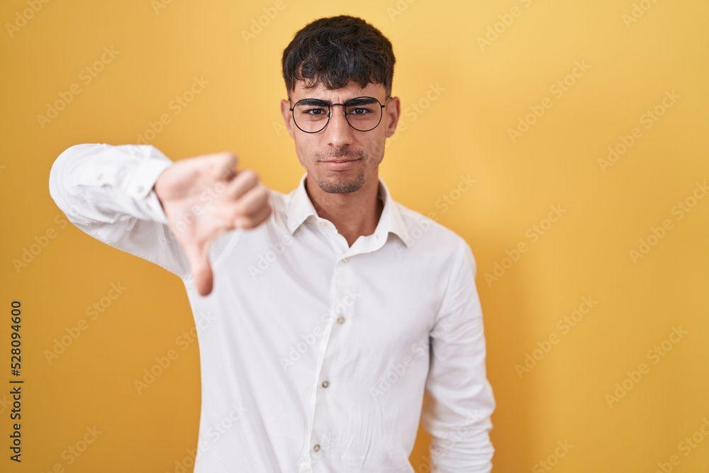Young hispanic man standing over yellow background looking unhappy and angry showing rejection and negative with thumbs down gesture. bad expression. Young hispanic man standing over yellow background looking unhappy and angry showing rejection and negative with thumbs down gesture. bad expression.