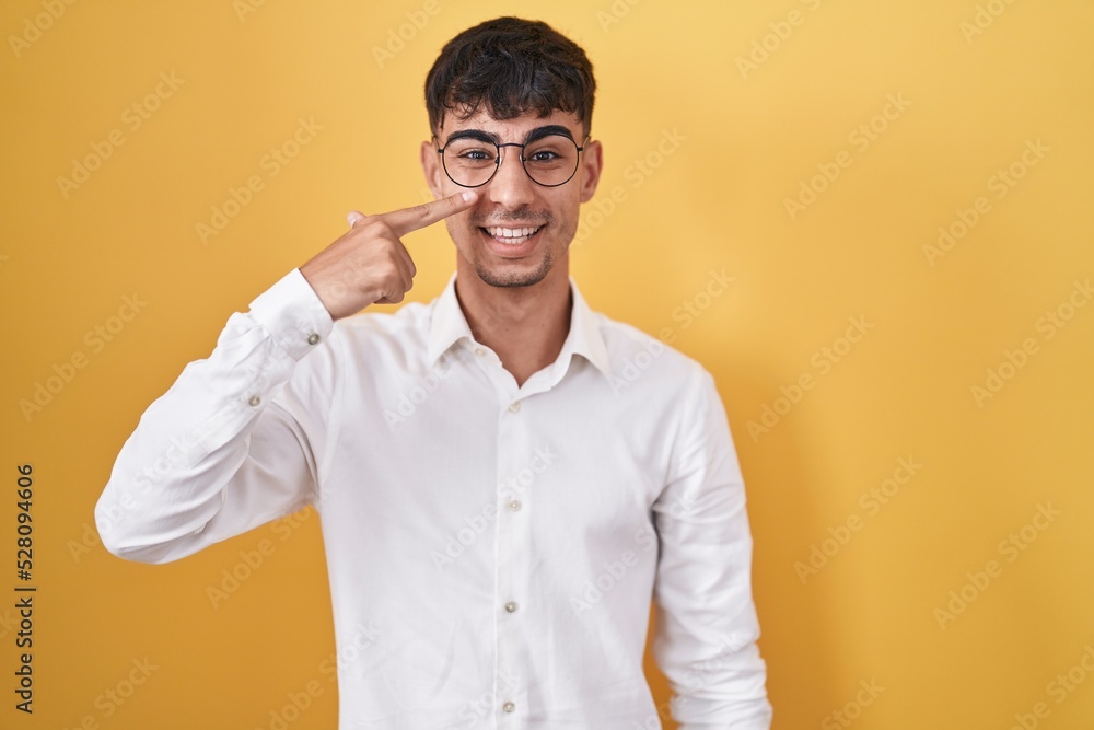 Young hispanic man standing over yellow background pointing with hand finger to face and nose, smiling cheerful. beauty concept Young hispanic man standing over yellow background pointing with hand finger to face and nose, smiling cheerful. beauty concept