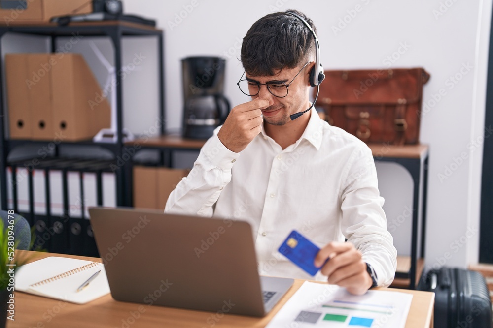 Young hispanic man working using computer laptop holding credit card smelling something stinky and disgusting, intolerable smell, holding breath with fingers on nose. bad smell Young hispanic man working using computer laptop holding credit card smelling something stinky and disgusting, intolerable smell, holding breath with fingers on nose. bad smell