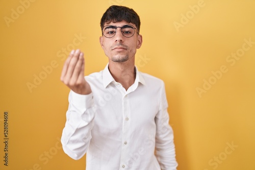 Young hispanic man standing over yellow background doing italian gesture with hand and fingers confident expression
