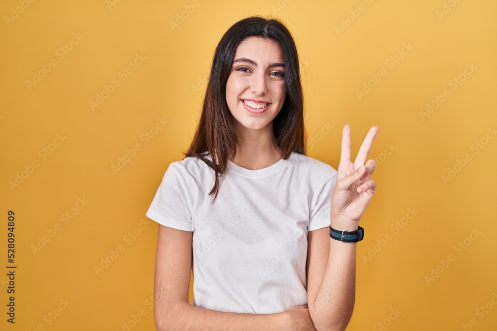 Young beautiful woman standing over yellow background smiling with happy face winking at the camera doing victory sign with fingers. number two. Young beautiful woman standing over yellow background smiling with happy face winking at the camera doing victory sign with fingers. number two.