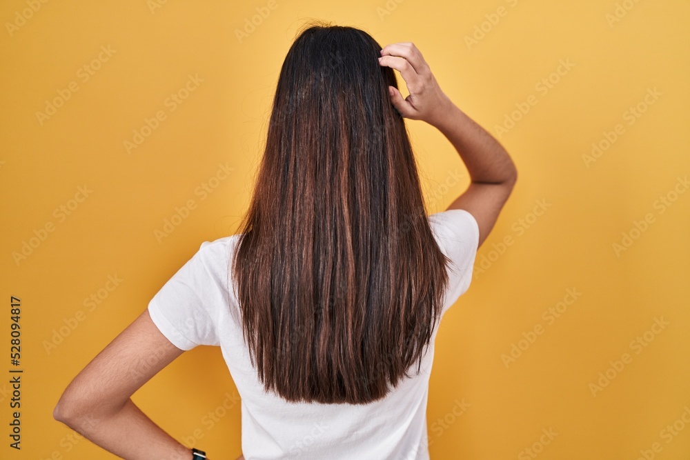 Young beautiful woman standing over yellow background backwards thinking about doubt with hand on head Young beautiful woman standing over yellow background backwards thinking about doubt with hand on head