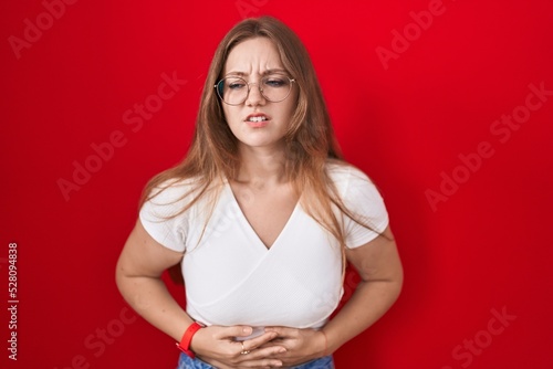 Young caucasian woman standing over red background with hand on stomach because indigestion, painful illness feeling unwell. ache concept.