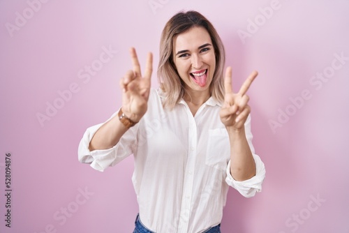 Young beautiful woman standing over pink background smiling with tongue out showing fingers of both hands doing victory sign. number two.