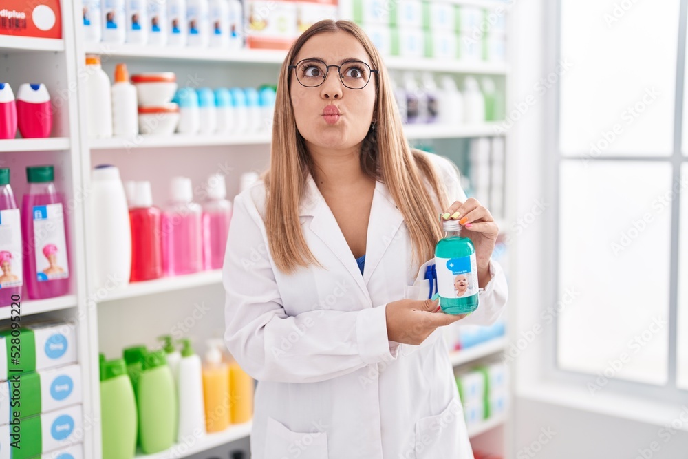 Young blonde woman working at pharmacy drugstore holding syrup making fish face with mouth and squinting eyes, crazy and comical.
