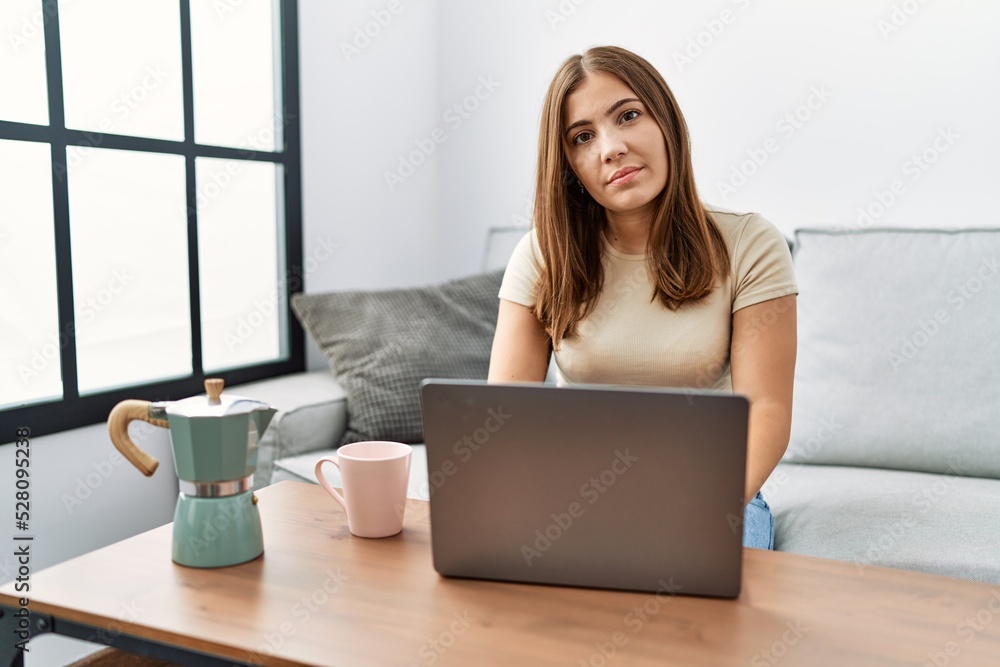 Young brunette woman using laptop at home drinking a cup of coffee relaxed with serious expression on face. simple and natural looking at the camera. Young brunette woman using laptop at home drinking a cup of coffee relaxed with serious expression on face. simple and natural looking at the camera.