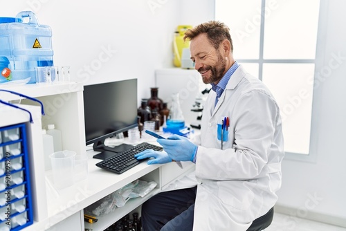 Middle age hispanic man wearing scientist uniform using smartphone at laboratory