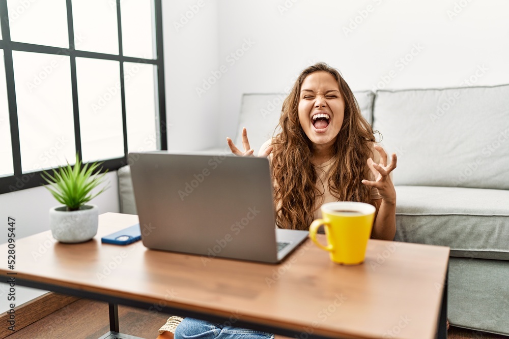 Beautiful hispanic woman using computer laptop at home crazy and mad shouting and yelling with aggressive expression and arms raised. frustration concept. Beautiful hispanic woman using computer laptop at home crazy and mad shouting and yelling with aggressive expression and arms raised. frustration concept.
