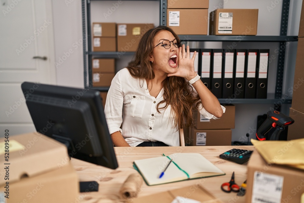 Young hispanic woman working at small business ecommerce shouting and screaming loud to side with hand on mouth. communication concept. Young hispanic woman working at small business ecommerce shouting and screaming loud to side with hand on mouth. communication concept.