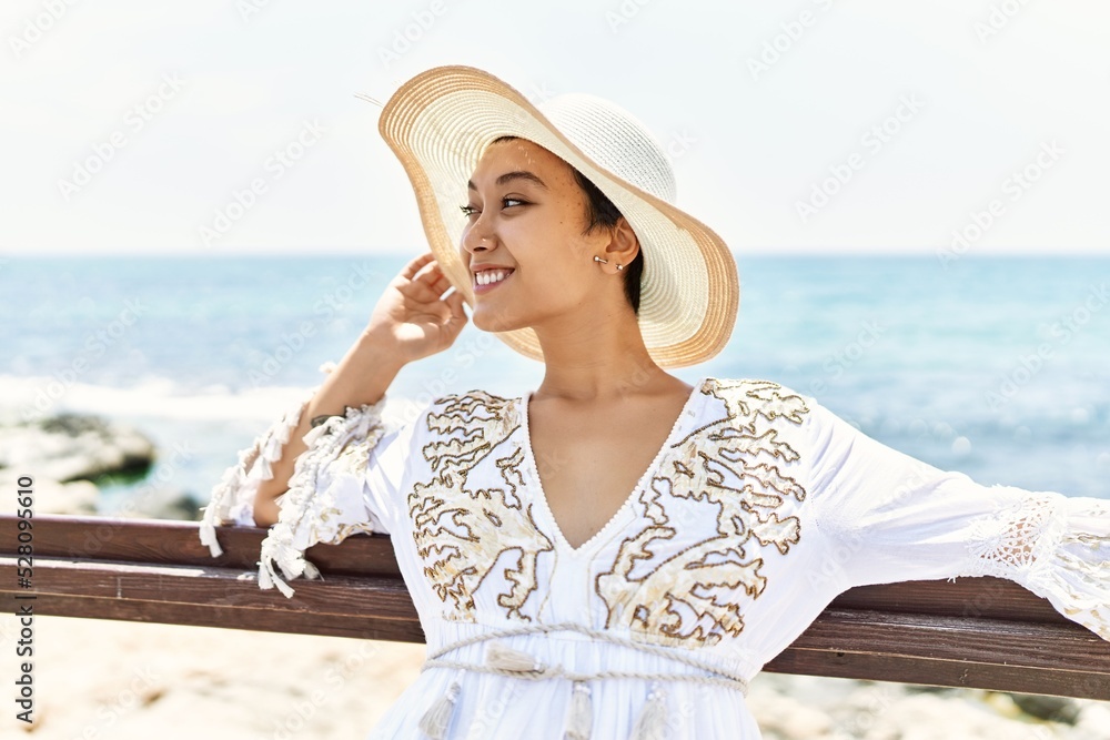 Young hispanic woman smiling confident wearing summer hat at seaside
