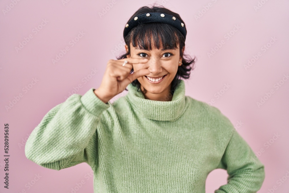 Young beautiful woman standing over pink background smiling and confident gesturing with hand doing small size sign with fingers looking and the camera. measure concept. Young beautiful woman standing over pink background smiling and confident gesturing with hand doing small size sign with fingers looking and the camera. measure concept.