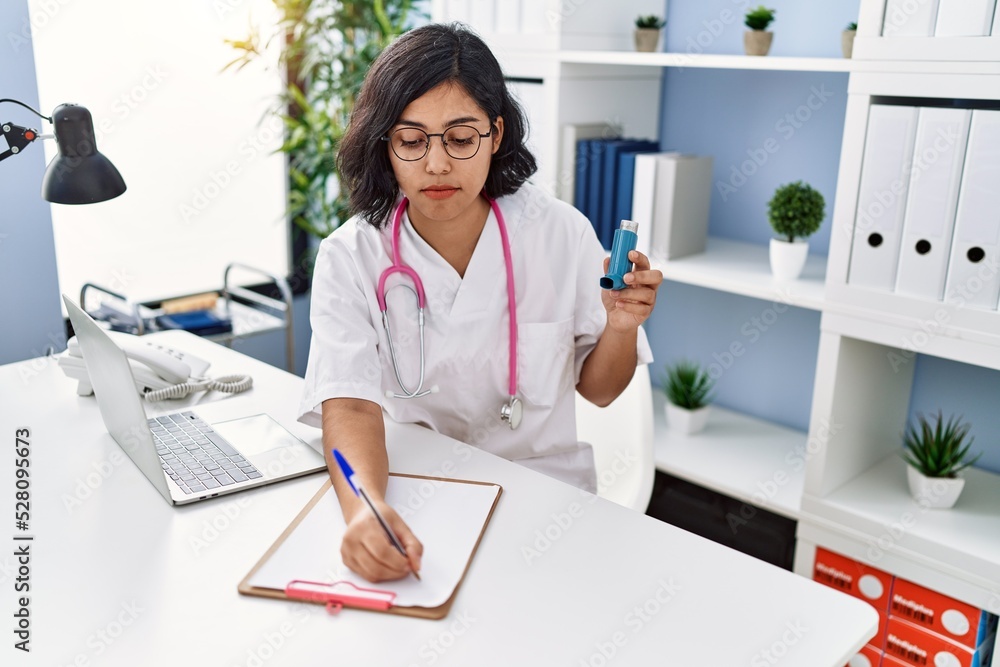 Young latin woman wearing doctor uniform holding inhaler writing on clipboard at clinic Young latin woman wearing doctor uniform holding inhaler writing on clipboard at clinic