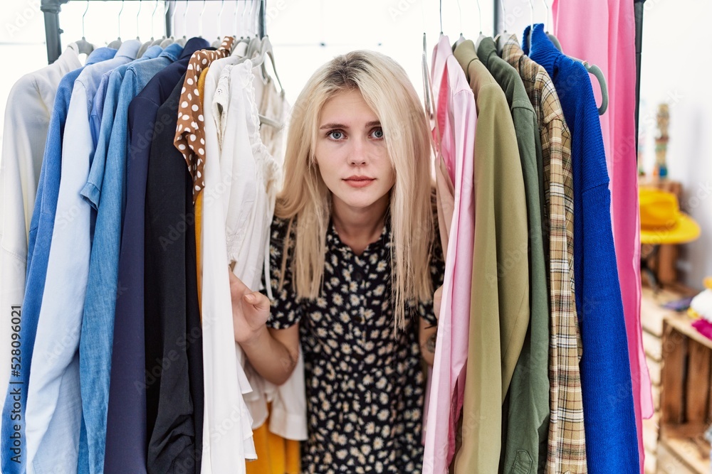 Young blonde woman searching clothes on clothing rack relaxed with serious expression on face. simple and natural looking at the camera.