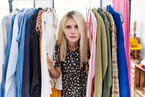 Young blonde woman searching clothes on clothing rack relaxed with serious expression on face. simple and natural looking at the camera.