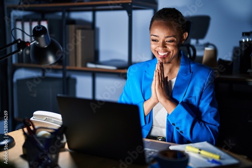 Beautiful african american woman working at the office at night praying with hands together asking for forgiveness smiling confident.
