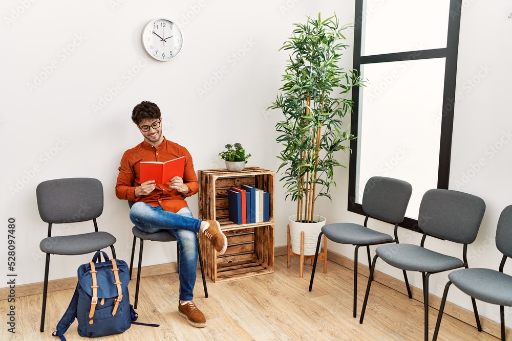 Fototapeta premium Young hispanic man smiling confident reading book at waiting room