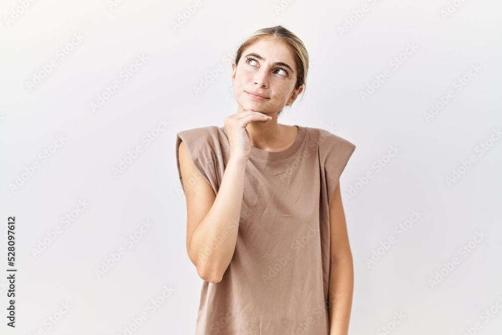 Young blonde woman standing over isolated background with hand on chin thinking about question, pensive expression. smiling with thoughtful face. doubt concept. Young blonde woman standing over isolated background with hand on chin thinking about question, pensive expression. smiling with thoughtful face. doubt concept.