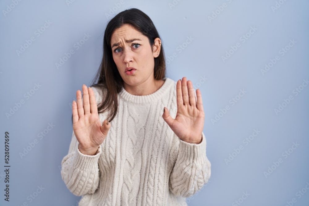 Young brunette woman standing over blue background moving away hands palms showing refusal and denial with afraid and disgusting expression. stop and forbidden.