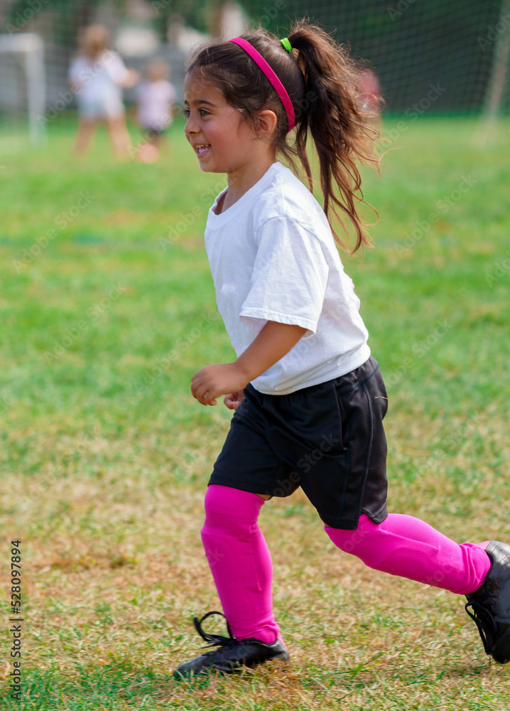 Young happy girl in generic peewee soccer uniform playing sports with her little teammates learning game outside on beautiful park field with green grass wearing pink uniform colors.
