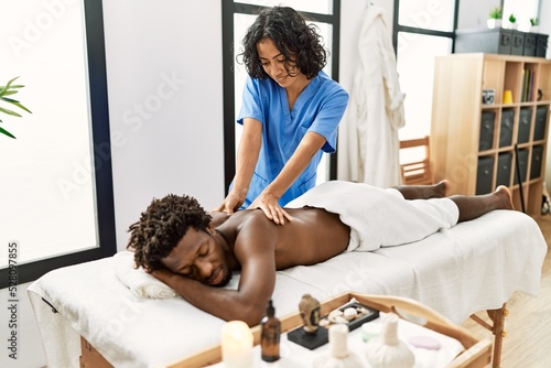 Young physiotherapist woman smiling happy giving back massage to african american man at the clinic.