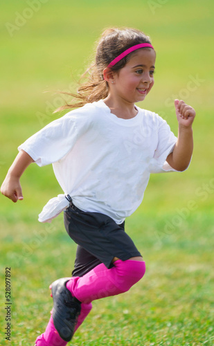 Excited young girl running on peewee soccer field learnings and playing football game with teammates outside in park on beautiful day time. Pink cleats and shin guards matching headband colors