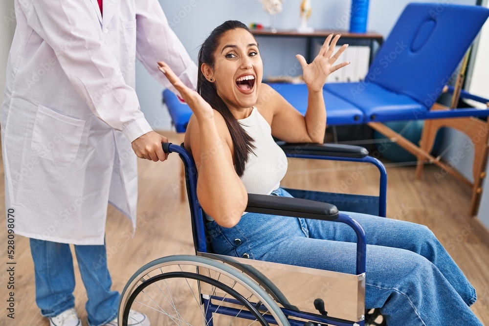 Obraz premium Young hispanic woman sitting on wheelchair at physiotherapy clinic celebrating victory with happy smile and winner expression with raised hands