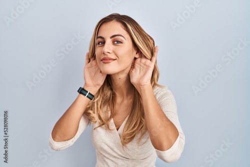 Young blonde woman standing over isolated background trying to hear both hands on ear gesture, curious for gossip. hearing problem, deaf