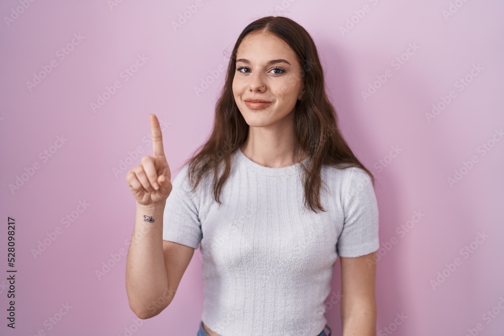 Young hispanic girl standing over pink background showing and pointing up with finger number one while smiling confident and happy. Young hispanic girl standing over pink background showing and pointing up with finger number one while smiling confident and happy.