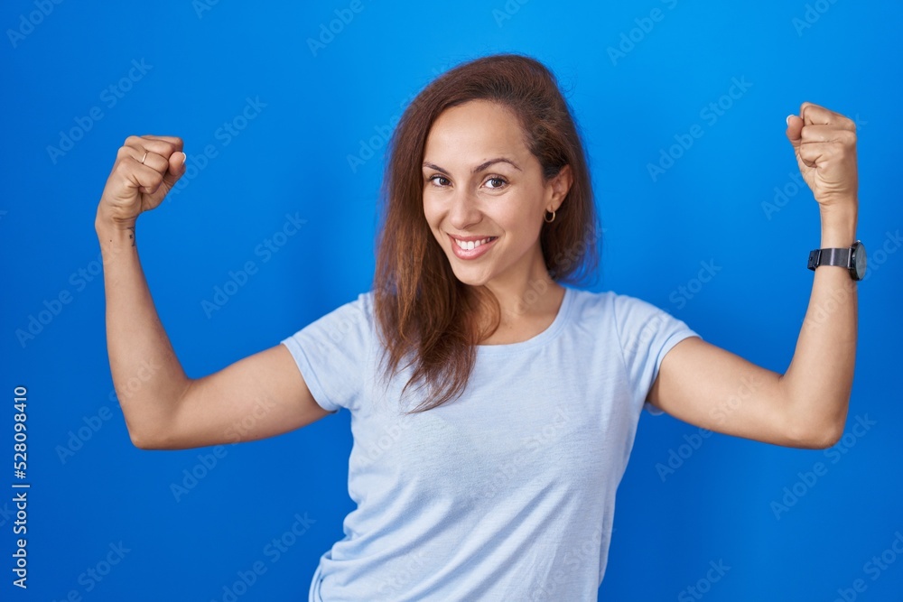 Fototapeta premium Brunette woman standing over blue background showing arms muscles smiling proud. fitness concept.