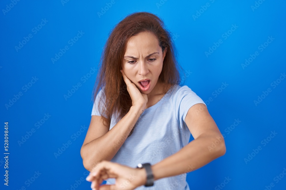 Brunette woman standing over blue background looking at the watch time worried, afraid of getting late Brunette woman standing over blue background looking at the watch time worried, afraid of getting late