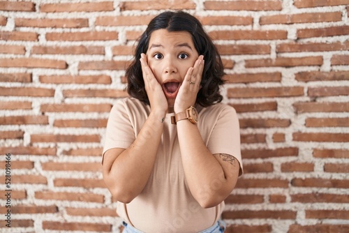 Young hispanic woman standing over bricks wall afraid and shocked, surprise and amazed expression with hands on face