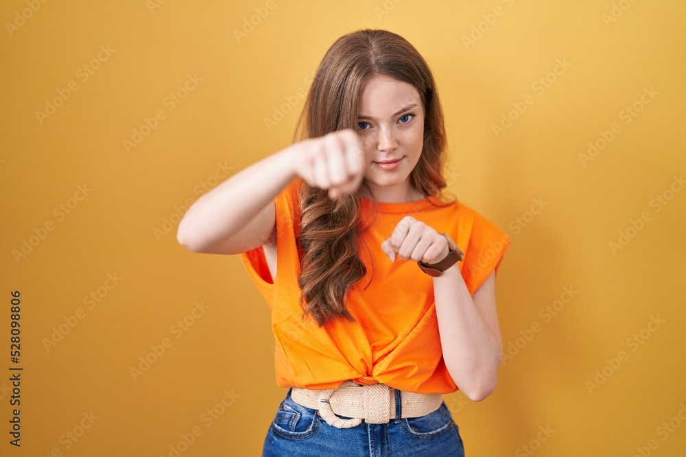 Caucasian woman standing over yellow background punching fist to fight, aggressive and angry attack, threat and violence